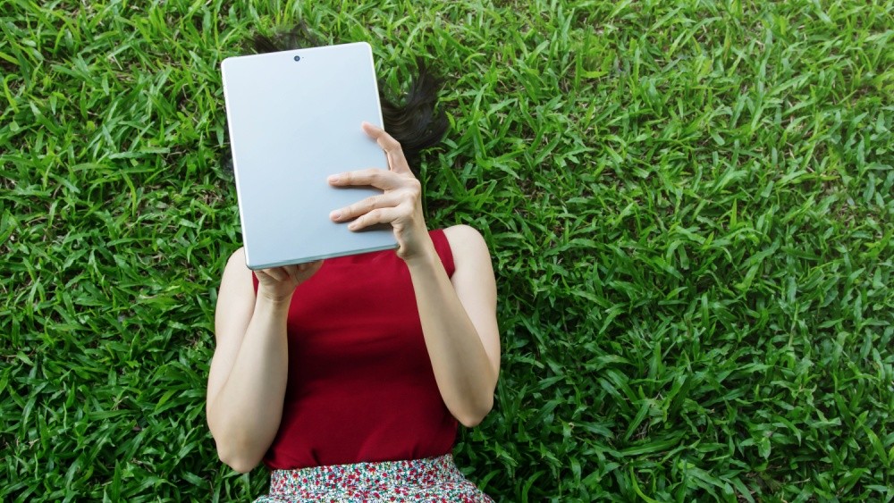 A woman lying in grass, looking at a tablet.
