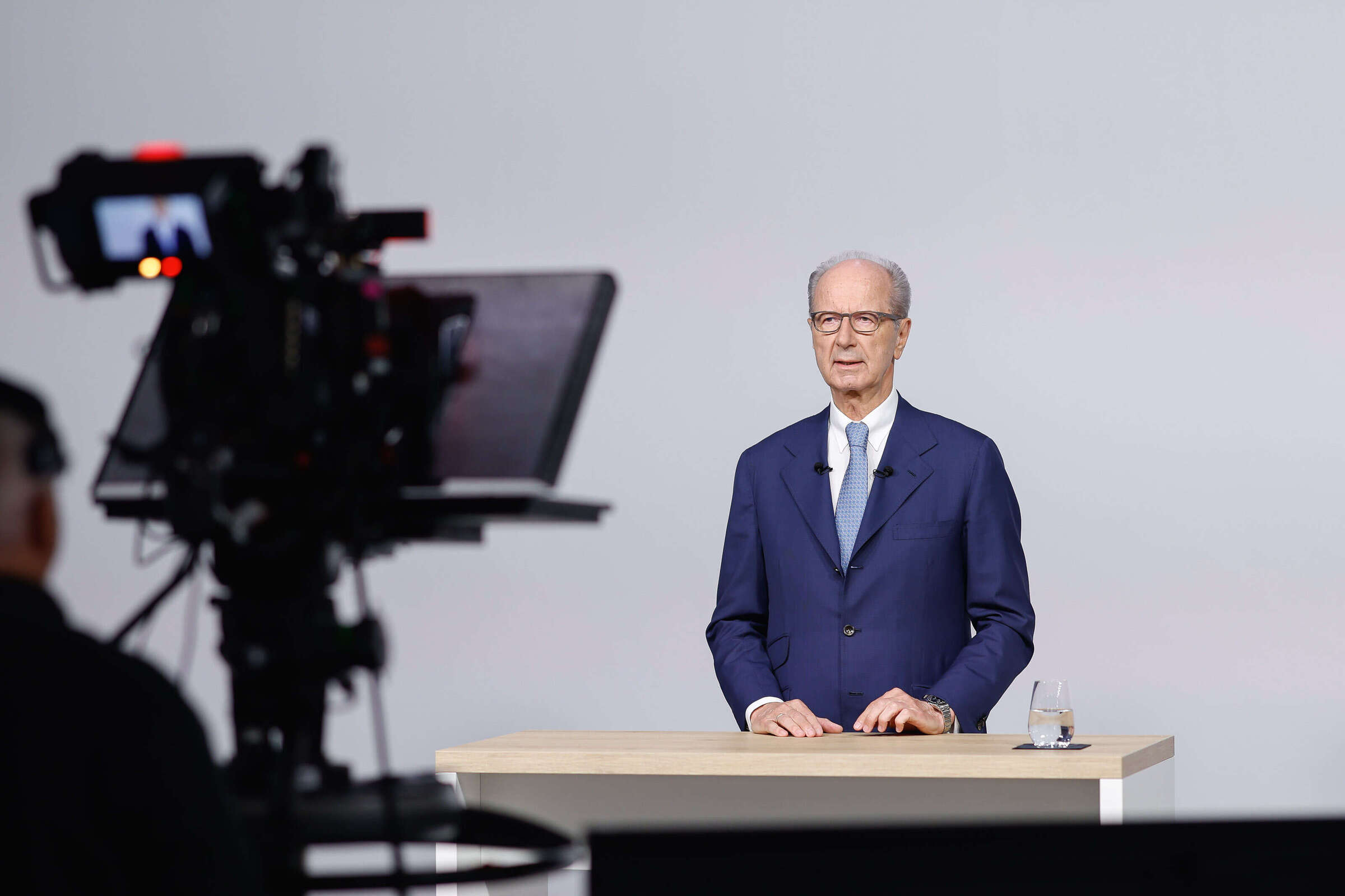 Hans Dieter P�tsch in a studio speaking behind a table to a camera; filming equipment in the foreground, a glass of water on the table.
