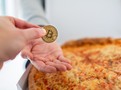 Ilustrační fotografie bitcoinu drženého v ruce nad pizzou, odkazující na Bitcoin Pizza Day a první známý nákup fyzického zboží za bitcoin.