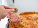Ilustrační fotografie bitcoinu drženého v ruce nad pizzou, odkazující na Bitcoin Pizza Day a první známý nákup fyzického zboží za bitcoin.
