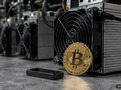 Symbolická bitcoinová mince a hardwarová peněženka před těžebním zařízením ASIC ilustrují volbu mezi těžbou a přímým nákupem Bitcoinu. Obrázek vytvořený pomocí AI.