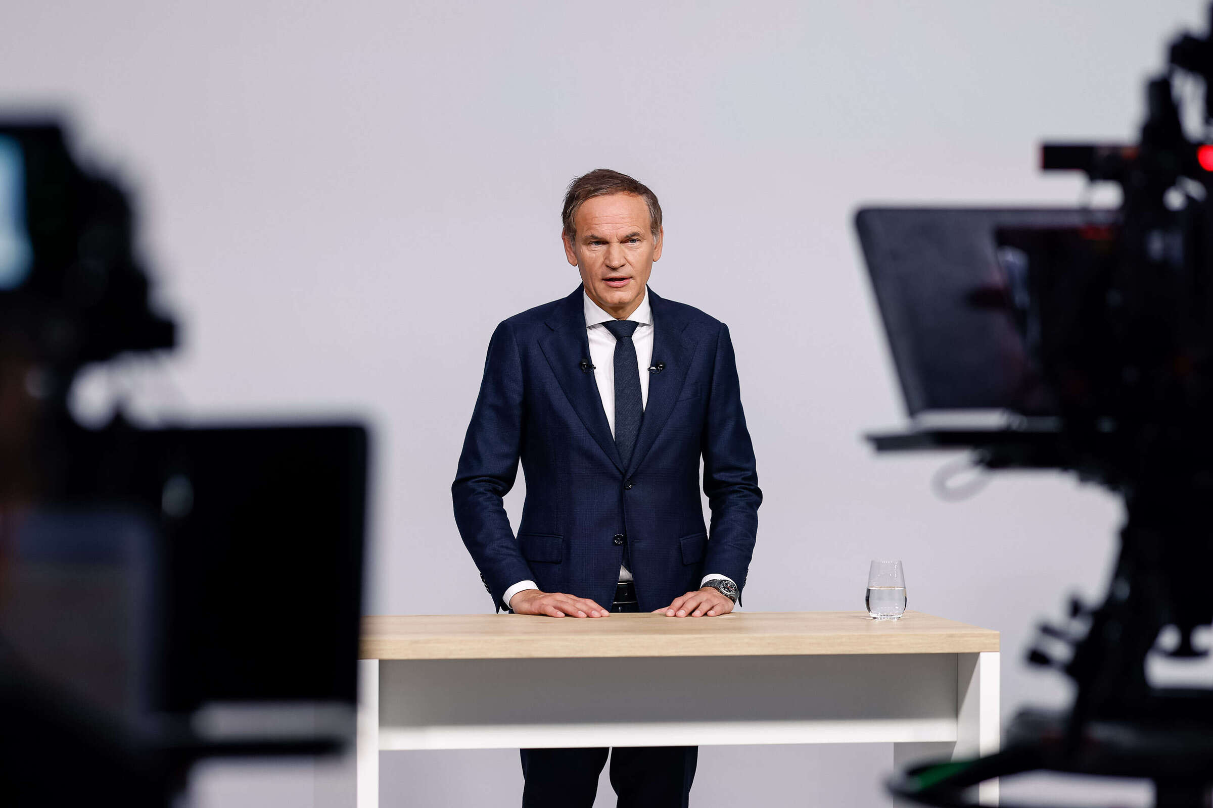 Oliver Blume in a blue suit at a light desk; blurred cameras in the foreground at the virtual VW Annual General Meeting.
